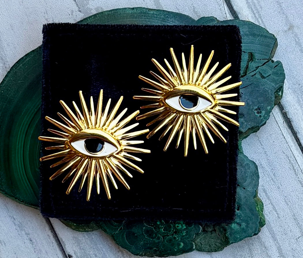 A pair of gold sunburst eye stud earrings displayed on a black velvet square, resting on a green agate slice over a white wood surface.